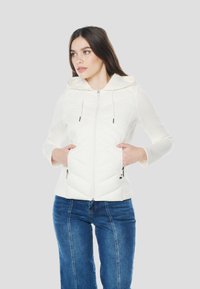 Fehér cipzáras kapucnis pulóver elülső részén cikk-cakk tűzéssel, húzózsinóros kapucnival és oldalzsebekkel. Kék farmernadrággal párosítva.