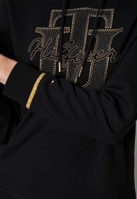 Sudadera negra con logo adornado con tachuelas doradas, cordones y un detalle dorado en la manga. Material de mezcla de algodón con una textura suave.