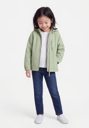 Παιδί που χαμογελάει και κοιτάζει προς τα κάτω, φοράει ανοιχτό πράσινο μπουφάν με κουκούλα, λευκό πουκάμισο, σκούρο μπλε τζιν και λευκά slip-on παπούτσια.