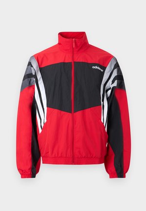 Punane ja must Adidas lukuga tuulekindel jakk kõrge kraega ja valgete triipudega varrukatel, laotatult valgel taustal.