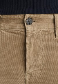 Beige ribfluweel broek met een geribde textuur, knoopsluiting met metalen accent en een zijzak. Bovenstuk is gemaakt van een geribde donkere stof.