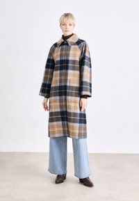 Plaid coat in brown, navy, and light blue tones, featuring a large collar, long sleeves, and two front pockets. Worn over wide-leg jeans.