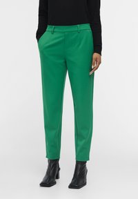 Pantalons verts à coupe ajustée, avec une texture lisse, des poches latérales et une taille, associés à des bottes à talons noires.