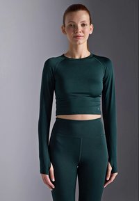 Ung kvinne med rødt hår iført en mørkegrønn langermet crop-top og matchende høyhøyde leggings mot en grå bakgrunn.