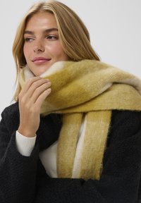 Gul og cremefarvet stribet tørklæde lavet af blødt, tekstureret stof, draperet omkring halsen. Båret over en mørk frakke og en lys sweater.