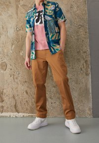 Koszulka z krótkim rękawem w kolorze niebieskim z kolorowymi wzorami geometrycznymi, w zestawieniu z różową koszulką i brązowymi spodniami; białe sneakersy dopełniają strój.