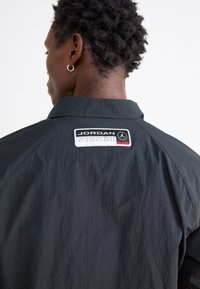 Μαύρο ελαφρύ μπουφάν με γιακά, διαθέτει ραμμένο ετικέτα με το "Jordan" και το λογότυπο. Ορατό υφασμάτινο σχέδιο και σχεδίαση με χαμηλούς ώμους.