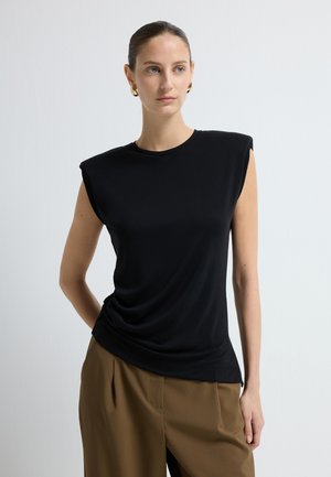 Musta hihaton paita, jossa on rakenteelliset olkapäät, laskostettu etuosa ja pyöreä pääntie, valmistettu sileästä kankaasta, yhdistetty vaaleanbeigeihin laskostettuihin housuihin.