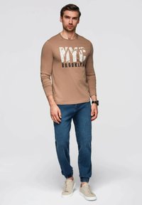 Camisa de manga larga beige con estampado "NYC BROOKLYN", combinada con joggers de mezclilla azules y zapatillas beige. Diseño sencillo, estilo ajustado.