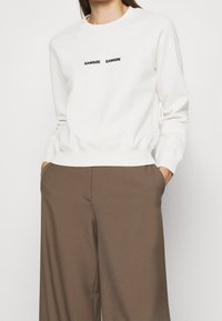 Sudadera blanca con cuello redondo, puños y dobladillo acanalados. Presenta el logotipo "SAMØE" en negro, combinado con pantalones anchos marrones.
