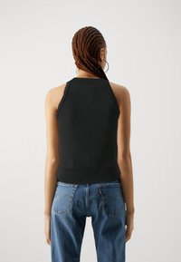 Svart strikket tanktopp med høy hals og ermeløs design, utfylt med en ribbet kant. Bæres med blå denimjeans.