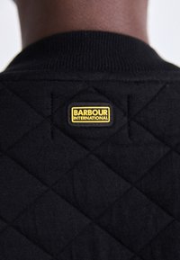 Tecido acolchoado preto com padrão de diamante, apresentando um rótulo retangular amarelo "BARBOUR INTERNATIONAL" na parte de trás do pescoço.