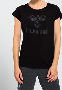 Černé bavlněné tričko s krátkým rukávem a kulatým výstřihem. Na přední straně se nachází lesklé černé logo "hummel" a grafický design.