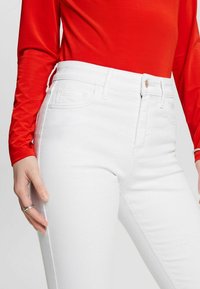 Jeans de mezclilla blanca de tiro alto con bolsillos delanteros, combinados con una blusa roja de manga larga. Textura suave y detalles de costura mínimos.