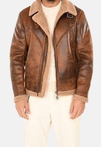 Mężczyzna w brązowej, postarzałej skórzanej kurtce z kołnierzem i mankietami z owczej wełny, beżowym swetrze oraz kremowych spodniach, stojący ze swobodnie opuszczonymi rękami.