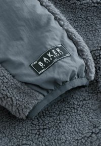 Szary polarowy materiał o teksturowanej powierzchni, z gładką szarością panelu i czarną metką z białym napisem "BAKER by TED BAKER."