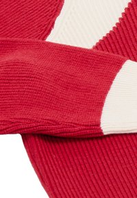Kötött pulóver piros alappal, fehér bordás mandzsettával. Az anyag enyhén vastag, ami hangsúlyozza a merész színkontrasztot és mintát.