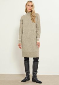 Abito lavorato a maglia beige con scollatura alta, polsini a costine e orlo. Indossato con stivali neri fino al ginocchio. Texture morbida e vestibilità ampia.