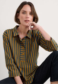 Camisa a rayas en color mostaza y azul, con cuello tipo camisero, botonera y un único bolsillo en el pecho. La tela parece suave.