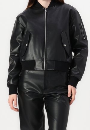 Vrouw draagt een zwarte bomberjas van nepleer met rits aan de voorkant en klepzakken, gecombineerd met bijpassende zwarte nepleren broek.