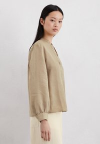 Beige Bluse mit langen Ärmeln, Mandarin-Kragen und Knopfdetail an den Manschetten, aus leichtem Stoff gefertigt, mit einem lockeren Schnitt.
