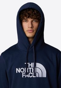 Sudadera con capucha azul marino que presenta un gran logo blanco con un diseño curvado. La tela parece texturizada y cuenta con un cordón en la capucha.