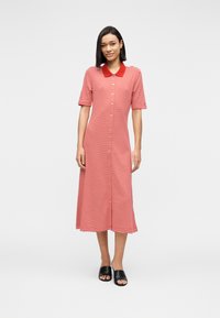 Γυναίκα όρθια, φορώντας κόκκινο και λευκό ριγέ φόρεμα με κόκκινο γιακά και μαύρα ανοιχτά υποδήματα τύπου slip-on, σε απλό φόντο.