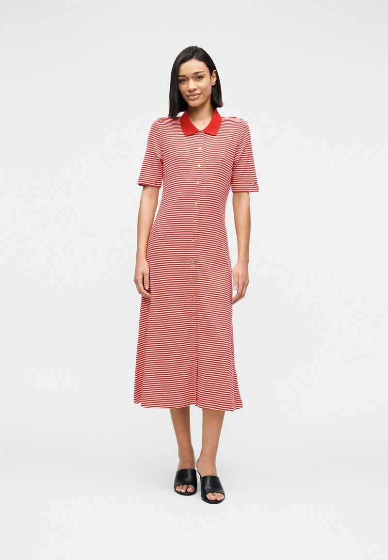 Γυναίκα όρθια, φορώντας κόκκινο και λευκό ριγέ φόρεμα με κόκκινο γιακά και μαύρα ανοιχτά υποδήματα τύπου slip-on, σε απλό φόντο.