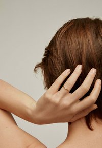 Anillo de oro con diseño delicado que presenta pequeñas piedras preciosas, llevado en una mano que se extiende hacia el cabello castaño hasta los hombros, contra un fondo neutro.