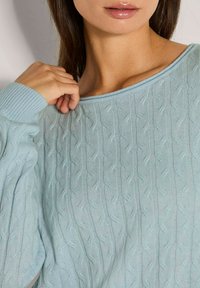 Eine Frau trägt einen hellblauen Kabelstrickpullover und hält mit einer Hand den Kragen, ihre Lippen glänzen und auf einer Seite sind dunkle Haare sichtbar.