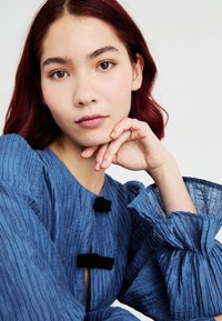 Top de tecido azul texturizado com mangas bufantes, detalhes em botão preto e um decote em forma de chave. Textura suave, design em camadas.