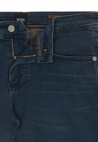 Jean en denim bleu foncé avec une fermeture éclair, des coutures bronze et un design classique à cinq poches. Présente une étiquette noire et un logo au niveau de la taille.