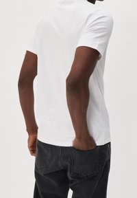 Herren-T-Shirt aus weißer Baumwolle mit kurzen Ärmeln und Rundhalsausschnitt, kombiniert mit dunkelgrauen Denim-Jeans. Der Stoff wirkt glatt und leger.