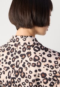 Korte bobkapsel gezien van achteren, draagt een beige blouse met een luipaardprint en een kleine ronde metalen tag bij de kraag.