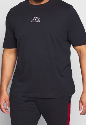 Mann trägt ein schwarzes Tommy Hilfiger T-Shirt mit kurzen Ärmeln und Logo auf der Brust, kombiniert mit schwarzen Hosen mit roten Seitenstreifen.