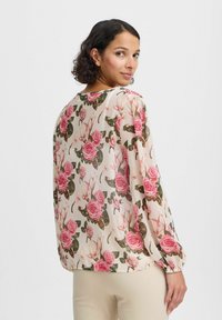 Durchsichtige Bluse mit beigefarbener Grundfarbe, die große rosa Rosen und grüne Blätter zeigt, mit langen Ärmeln und elastischen Bündchen sowie einem runden Halsausschnitt.