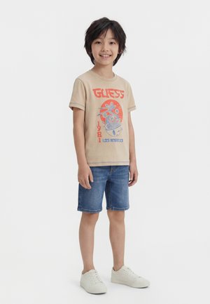 Jeune garçon debout et souriant, portant un t-shirt graphique beige, un short en jean et des baskets blanches sur un fond blanc uni.