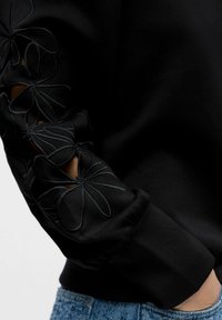 Svart genser med blomsterbroderi på ermene, med utskjæringer som viser hud. Tettsittende mansjetter og myk tekstur.