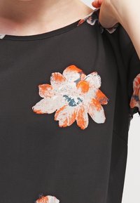 Černá blůza s velkými, živými květinovými vzory v oranžové a bílé barvě se modrým středem. Měkká, splývavá látka a kulatý výstřih.