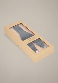 Czarne szelki w przezroczystym pudełku z zewnętrzną warstwą z papieru kraft i srebrnymi metalowymi klipsami. Na tkaninie znajduje się okrągłe logo.