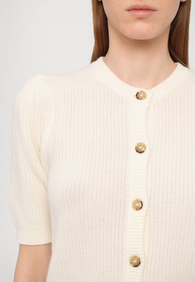 Cream-colored short-sleeve cardigan with a textured knit pattern, featuring a buttoned front and round neckline, adorned with circular buttons.