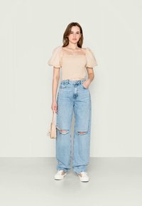 Beige puffærmet bluse over lyseblå ripped wide-leg jeans, suppleret med hvide sneakers og en lille beige håndtaske. Glat tekstur overordnet.