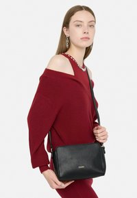 Sort læder håndtaske med en struktureret finish, udstyret med en lang rem og subtile guld-logo detaljer. Modellen bærer en rød off-shoulder sweater.