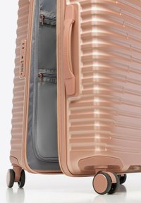 Valise rigide en or rose avec surface texturée, fermeture éclair, intérieur gris et roulettes doubles pour la mobilité. Dispose d'une poignée latérale et d'un verrou.