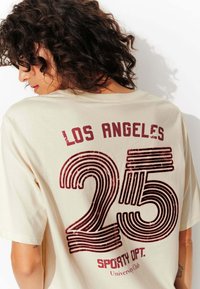 T-shirt crème avec le texte bordeaux "LOS ANGELES" et "SPORTY OPT." et un design audacieux rayé "25" au dos.