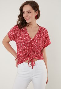 Rød blomsterbluse med korte ærmer, V-udskæring og samlede detaljer ved taljen bundet med et bånd. Parret med hvide jeans.