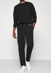 Černá mikina s bílým límcem a ladící kalhoty, které mají stahovací pas a boční panely, nošena se bílými teniskami.