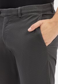 Zbliżenie na dłoń wsuniętą w przednią kieszeń ciemnoszarych spodni o teksturze, noszonych z czarną koszulą.