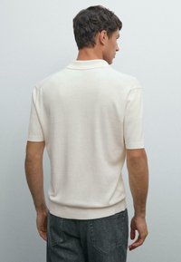Man med kort mörkt hår iförd en krämfärgad kortärmad polo-skjorta och mörkgrå jeans, stående vänd mot en ljusgrå vägg.