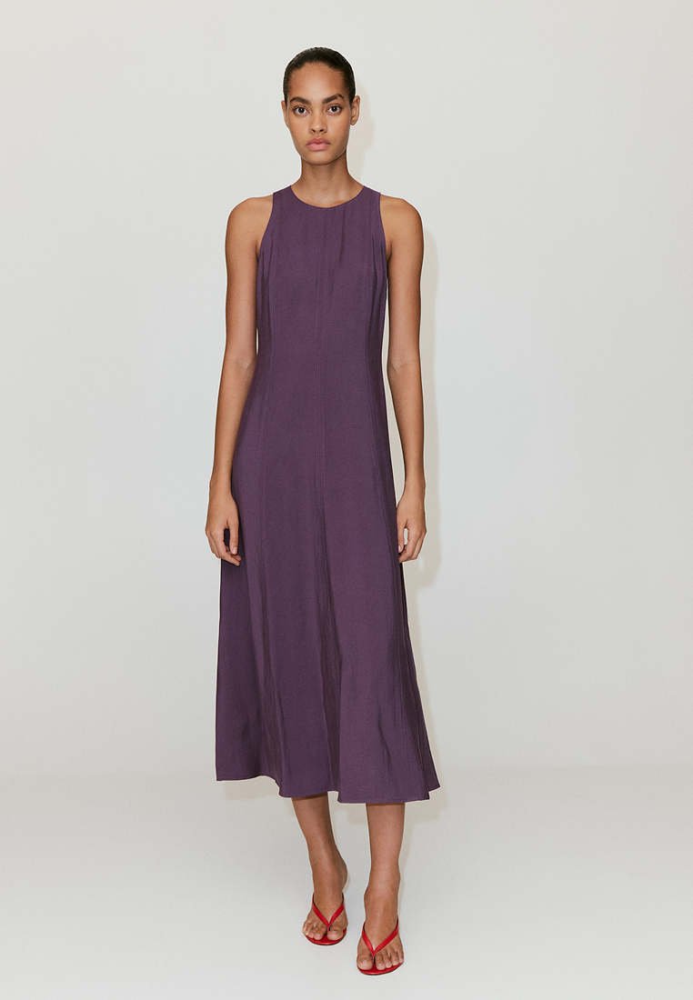 Femme debout portant une robe midi sans manches violet foncé et des sandales rouges à talons hauts sur un fond clair uni.